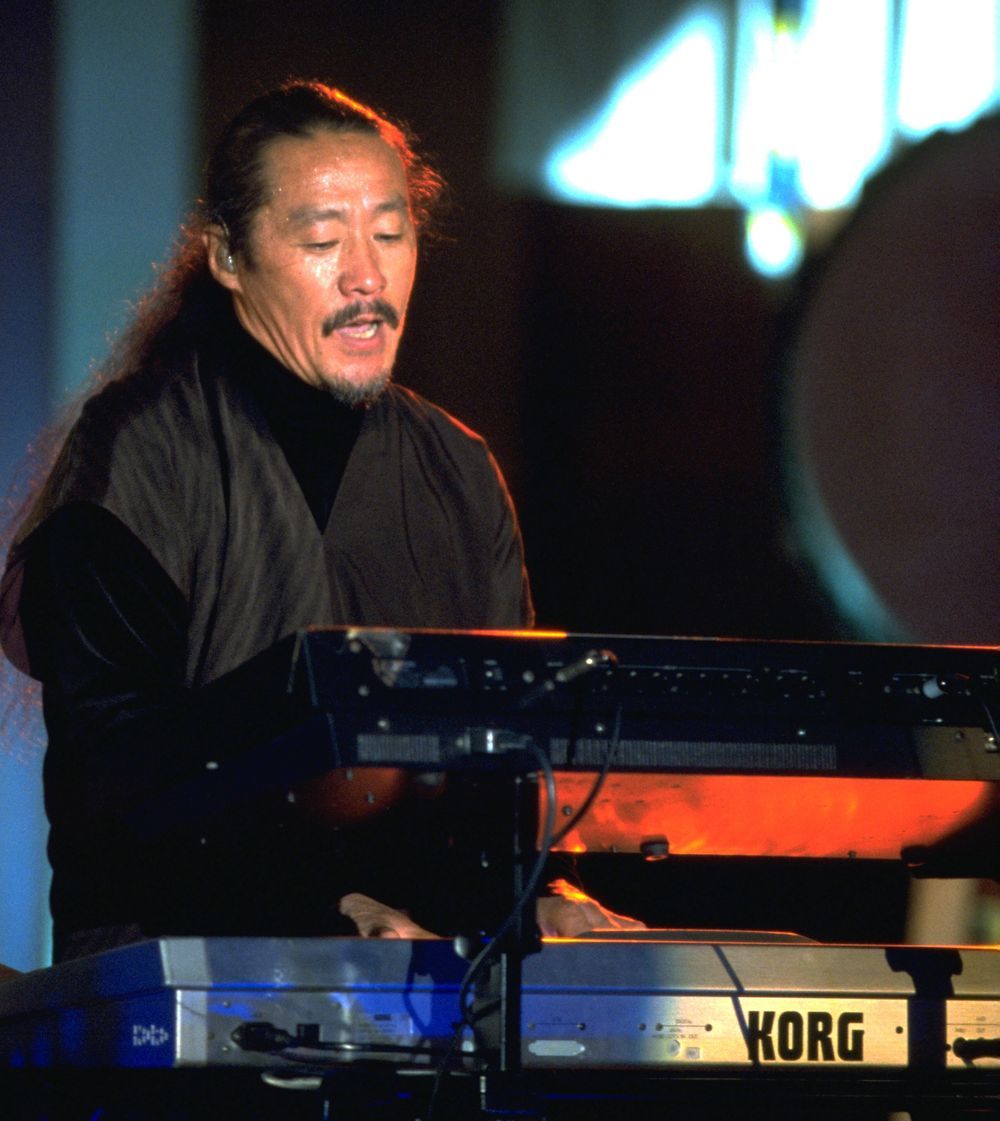 Kitaro on stage in black, long hair tied back, playing a Korg keyboard