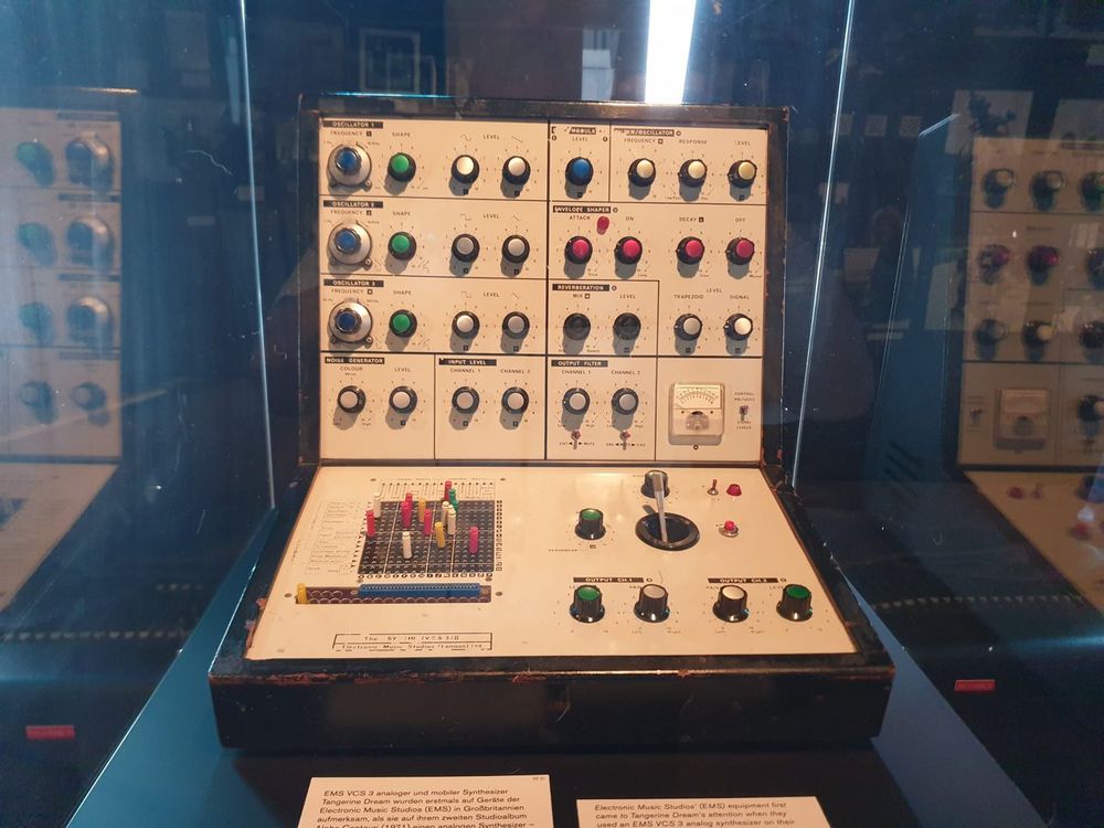 An EMS Synthi VCS 3 in a museum case. Rows of knobs fill the raised upper panel, and coloured pins stand in the matrix on the lower left