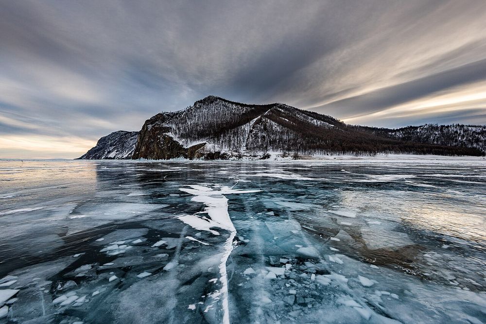 A frozen lake, clear cracked ice in front of a snow-covered rocky hill