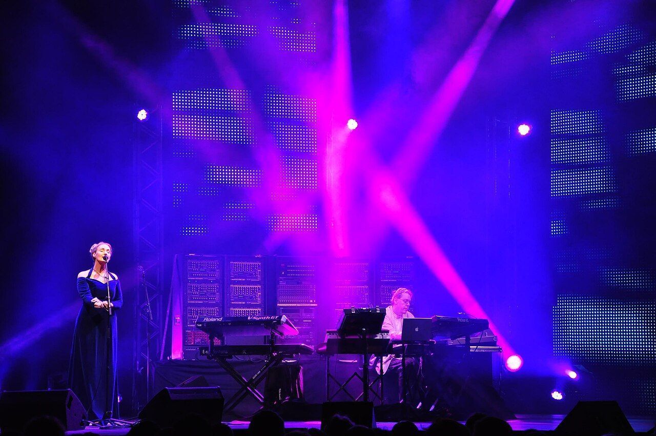 Klaus Schulze on stage behind a wall of Moog modules, with Lisa Gerrard singing, in blue and purple light