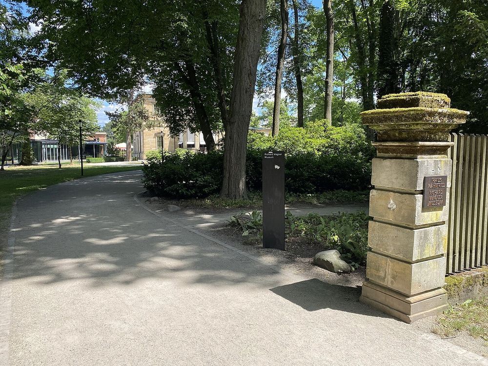 The garden entrance to Haus Wahnfried in Bayreuth. A mossy stone gatepost carries a bronze plaque reading Haus Wahnfried Richard Wagner Museum, and the house stands at the end of a path through the trees