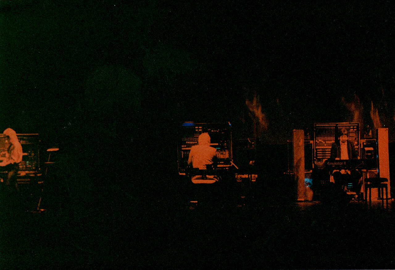 Tangerine Dream on a dark stage in the 1980s, three figures at banks of equipment, an Emulator II visible